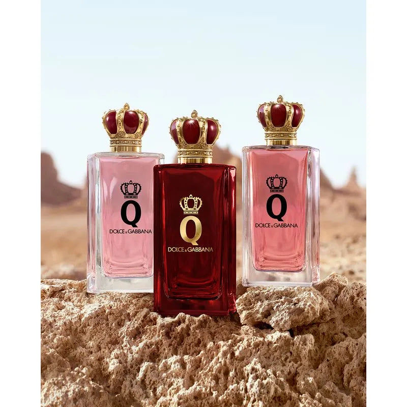 Three Dolce & Gabbana Q perfume bottles arranged on sandy ground with a desert landscape and pale sky in the background. The center bottle is dark red with a gold crown and black Q logo, while the two side bottles are light pink with matching crowns and Q logos.