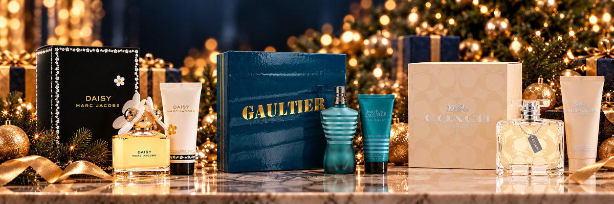 A luxury fragrance gift set arranged on a marble surface in front of a festive Christmas tree with warm golden lights. The set includes a dark blue GAULTIER box, a black DAISY MARC JACOBS box, a blue striped DAISY MARC JACOBS perfume bottle, a blue DAISY MARC JACOBS cologne bottle, a beige DAISY MARC JACOBS box, a white tube, and a clear bottle with a gold label.