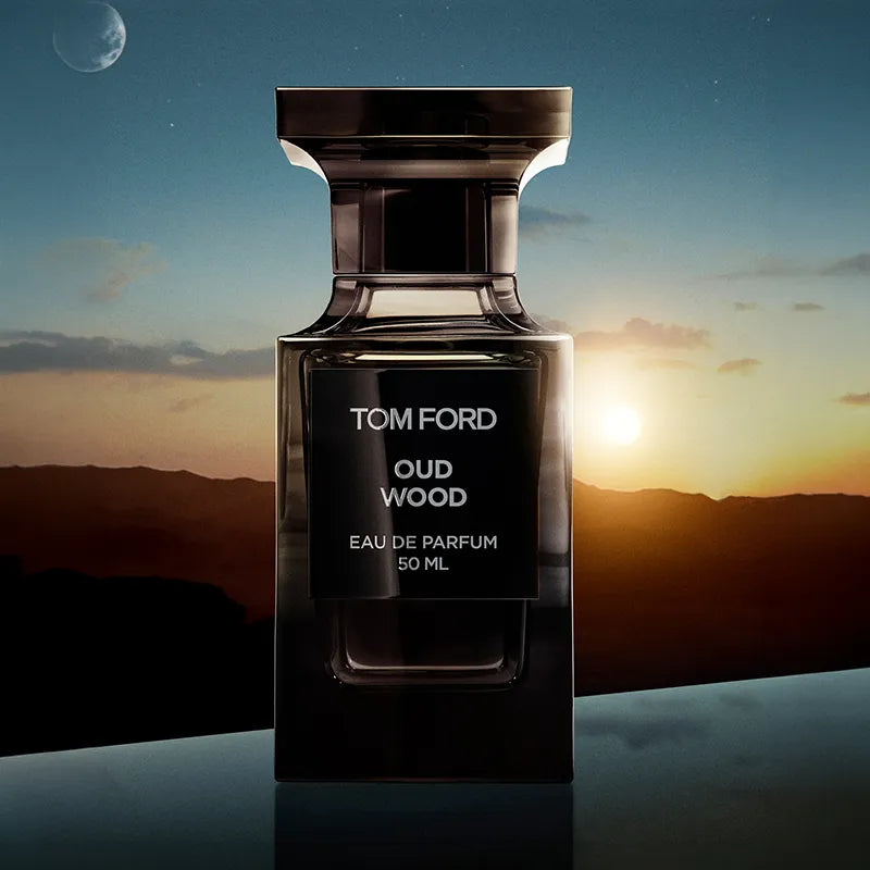 A 50 ml bottle of Tom Ford Oud Wood Eau de Parfum, presented in a dark, reflective glass bottle with a black label. The background is a dramatic sunset landscape with mountains, a glowing sun, and a moon in the sky.